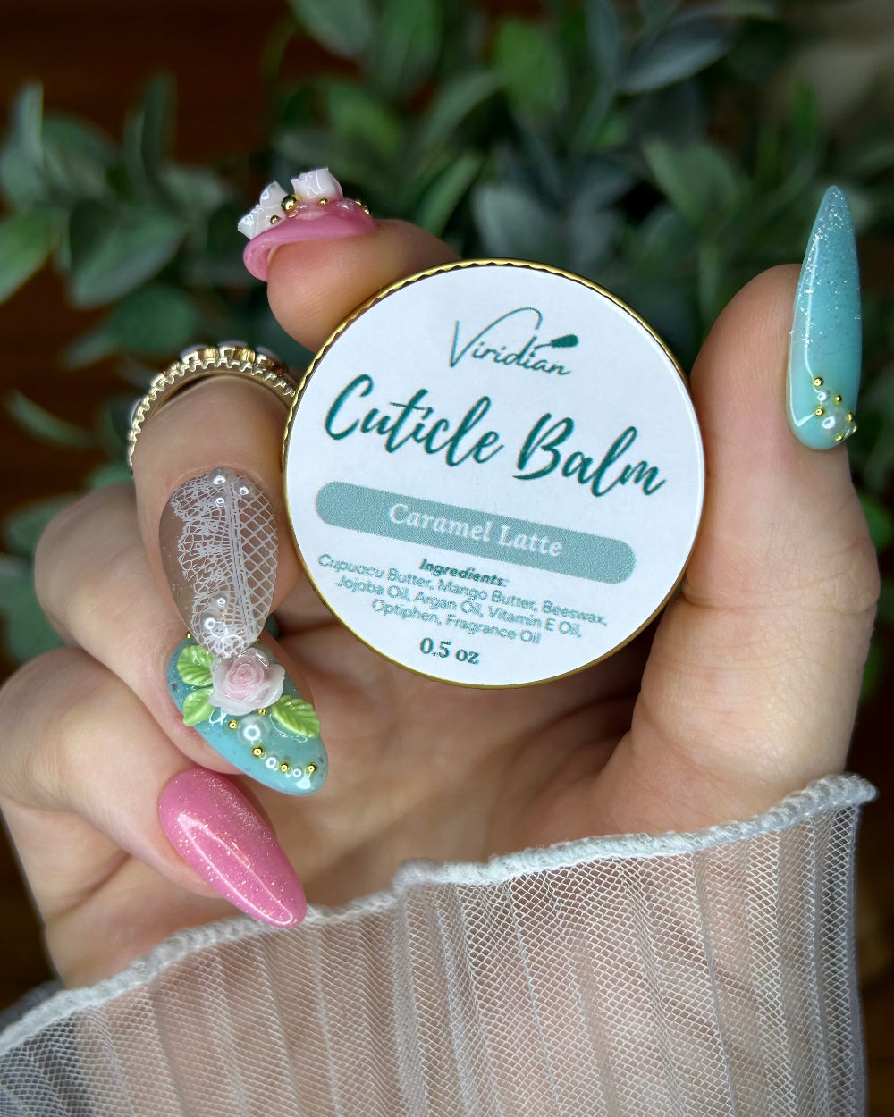 Hand holding a container of Viridian Cuticle Balm with a blurred green plant background