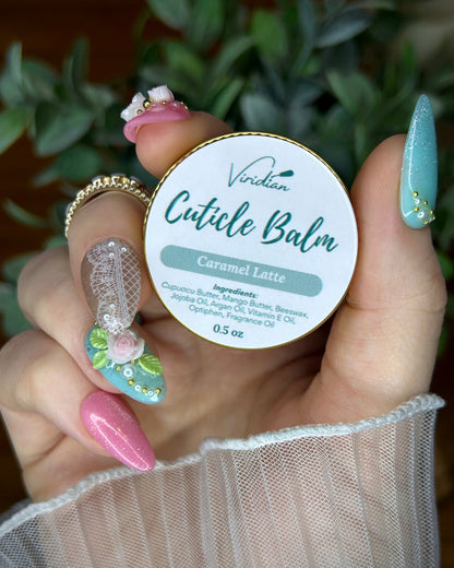 Hand holding a container of Viridian Cuticle Balm with a blurred green plant background