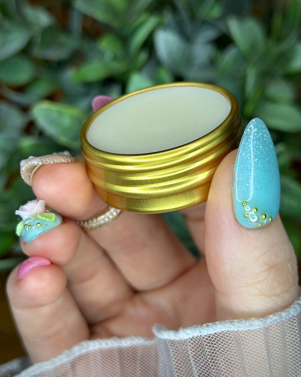 Gold ring with a white stone held by a hand with light blue nail polish against a green leafy background