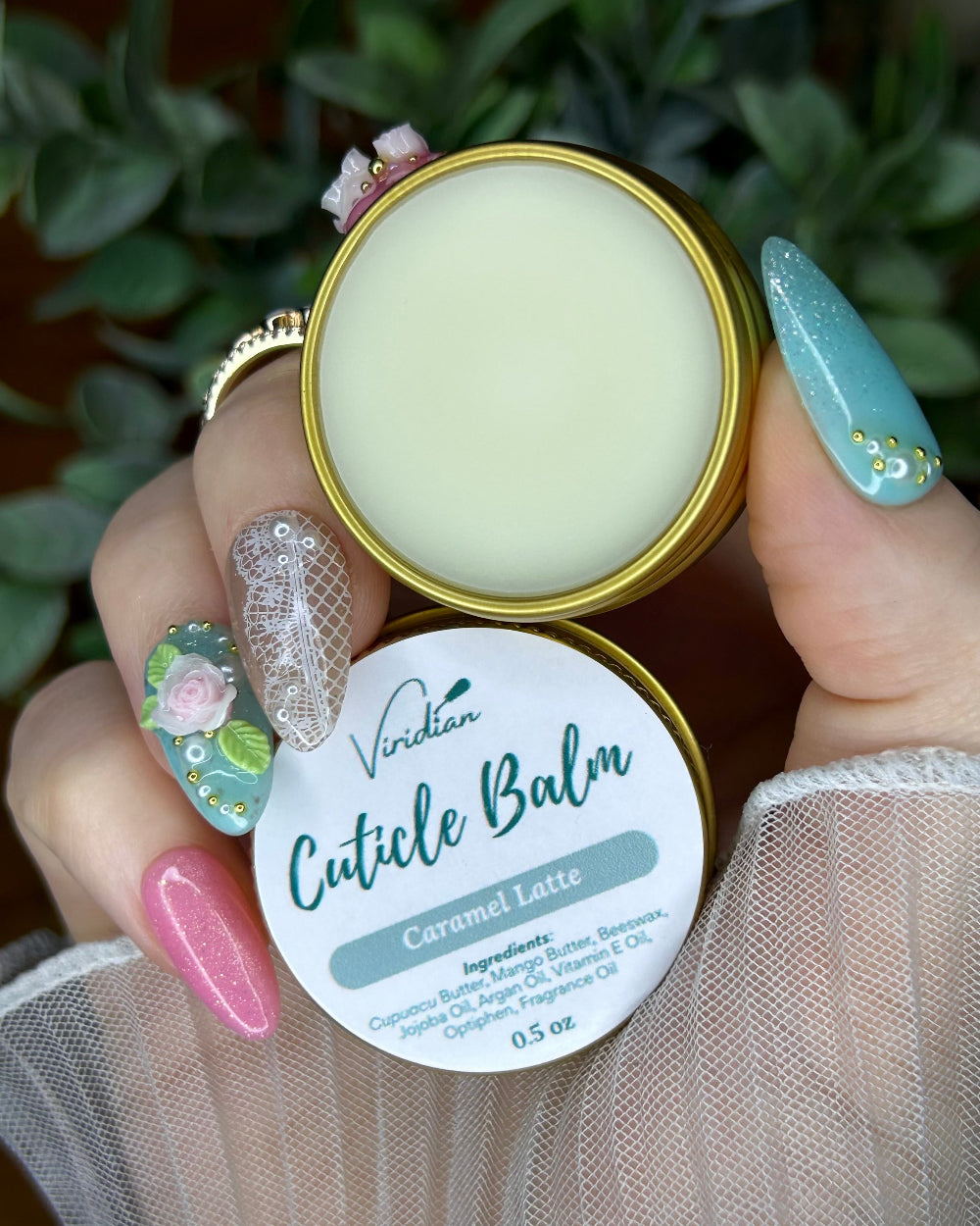 Hand holding a container of Viridian Cuticle Balm with a green plant in the background